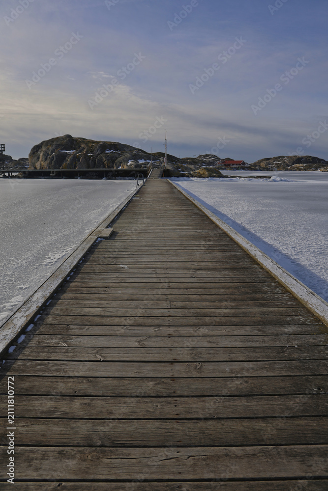 Fototapeta premium Symmetrical view of a wooden pier with rope railings leading across the ice in Skrhamn. A cold winter day in the Swedish archipelago with dramatic light and frozen wate