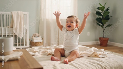 An adorable baby sitting on a bed with arms raised in joy, laughing happily in a bright nursery with a crib and a plant in the background.