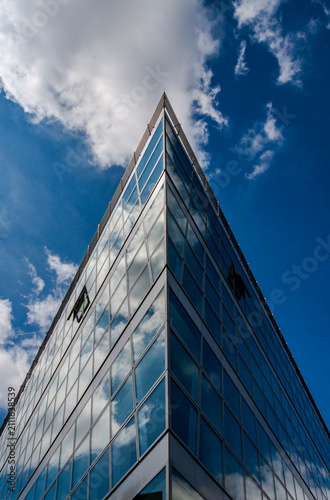 Close-up of the corner of a modern office building with a glass facade. Bright sunshine reflects white clouds in the windows, emphasizing clean architectural lines and contemporary design.