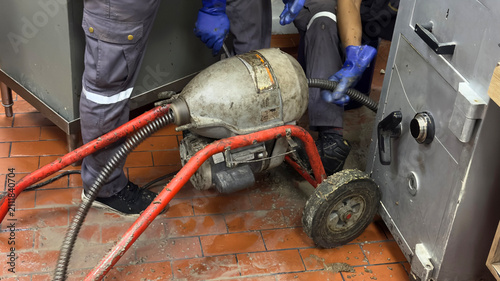 Konstfotografi Professional plumbers operating an electric drain cleaner machine, feeding a flexible steel auger cable to clear a clogged pipe on a commercial kitchen floor