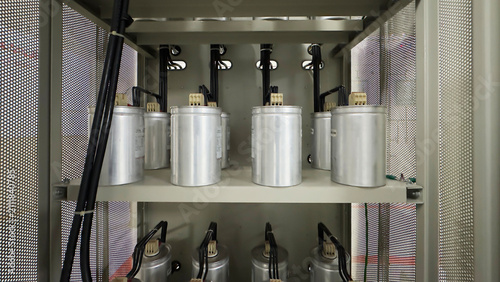 Row of industrial power factor capacitors inside electrical control panel cabinet with heavy wiring and cable connections.