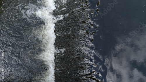 Photos Top down view of flowing water streaming over a small dam or weir threshold creating white foam, splashes, and dark water reflections