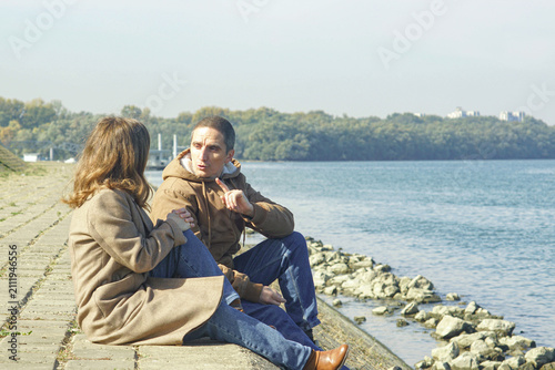 Quadro su tela Man raises one finger while speaking seated beside woman on river embankment