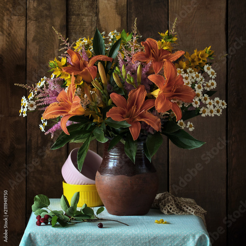 wildflower bouquet with orange daylilies sits in a ceramic vase next to stacked bowls. Floral decoration, botanical arrangement, and rustic still life concept