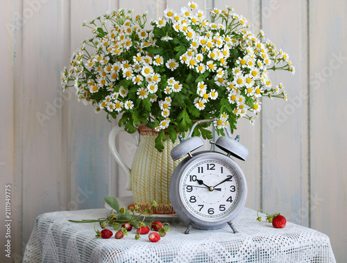 Chamomile bouquet in a ceramic pitcher stands next to a vintage alarm clock on a lace tablecloth. Still life decoration