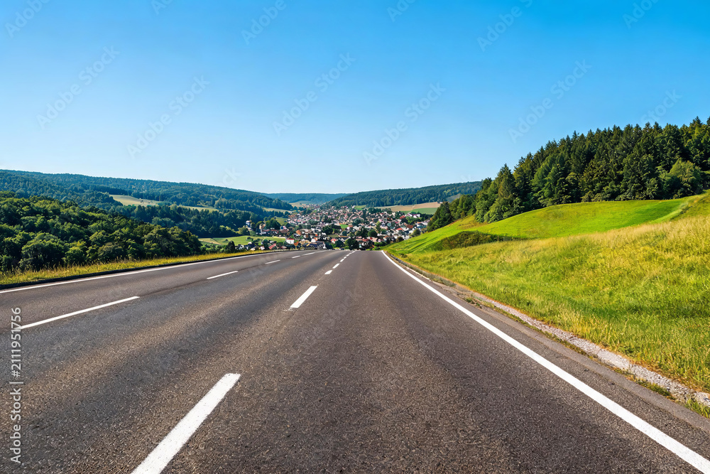 Obraz premium A two-lane asphalt highway stretches toward a small town nestled in rolling green hills under a clear blue sky.
