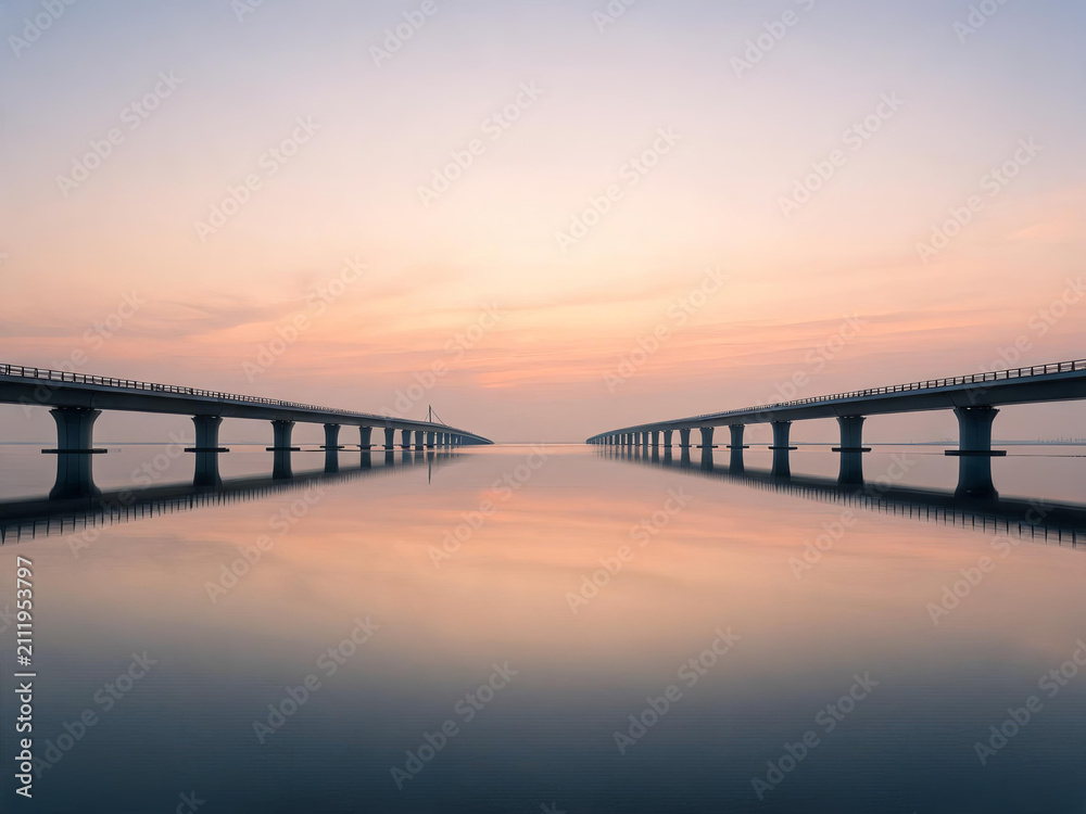 Obraz premium Two parallel bridges extend over calm reflective water, framed by a soft pastel sunset sky creating a tranquil symmetrical scene.