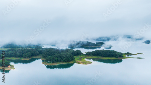 A tranquil aerial view of a misty lake with forested islands, calm water reflecting the green land, shrouded in soft fog.