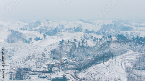 A wintry landscape of rolling snow-covered hills, with scattered trees and a small rural settlement nestled in the hilly terrain under hazy skies.