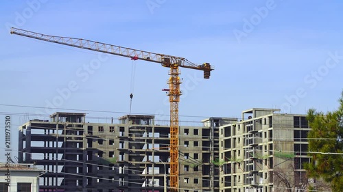 Video of a construction site in which work is underway to erect a multi-storey building using a yellow tower crane. The hook and free berths are lifted, the slinger gives hints to the crane operator