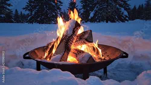 A warm campfire burns in a metal pit during a snowy night