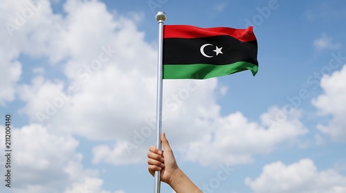 Person holding Libyan flag waving on silver flagpole against bright blue sky with white clouds
