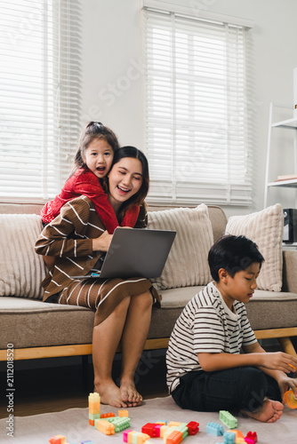 Asian mother working on laptop on couch while children play with building blocks at home