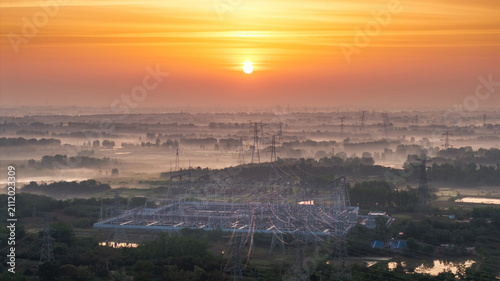 Foto A sunrise illuminates a misty rural landscape with an electrical substation and power pylons, blending natural and man-made elements