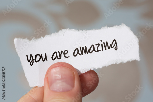 Romantic handwritten text on a paper note. A man is holding a piece of paper with text to her or him.. Text says you are amazing.