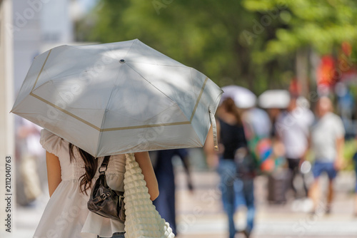 Rear view of a woman carrying a parasol on a city street in dappled light during summer heat with a blurred pedestrian crowd background.