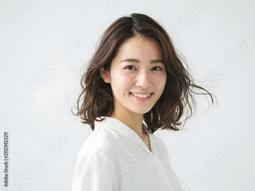 Smiling young japanese woman with dark wavy hair looking at the camera wearing a white shirt against a solid light grey studio background