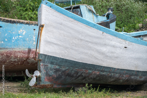 Canvastavla the weathered white and blue stern of an old boat, complete with a visible propeller and rudder, sits grounded on a grassy patch in an outdoor boatyard