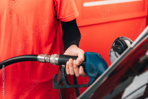 a close-up view of a person refueling a vehicle. The focus is on the individual's hand gripping a blue-handled fuel pump nozzle that is inserted into the open fuel tank of a dark car.