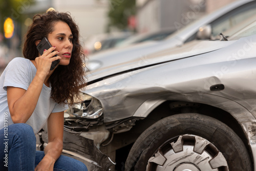 фотографія Young woman making a phone call to her insurance company after a traffic accident while inspecting damage to her car