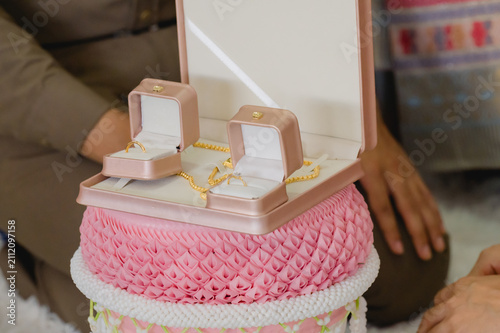 Obraz na plátně Two gold wedding rings inside pink boxes rest beside gold necklace inside traditional Thai ceremonial tray, representing dowry presented inside marriage ritual, couple sitting background indoors