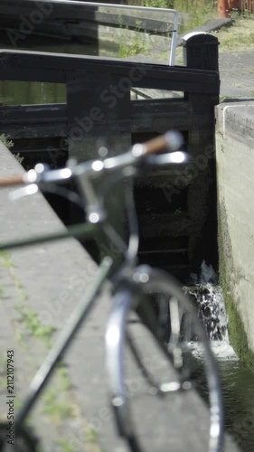 Bicycle by Grand Union Canal Lock Gate in Birmingham.
A vintage city bicycle rests in soft focus in the foreground as water flows through traditional wooden canal lock gate on the Grand Union Canal. 