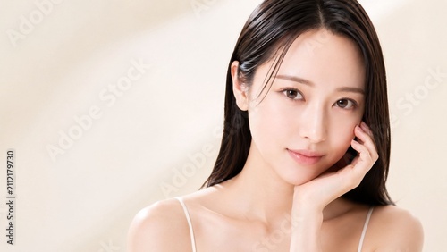 柔らかなベージュ背景を背に、片手を頬に添えてカメラを見つめるロングヘアの若い女性を写した上品なスキンケア美容ポートレート