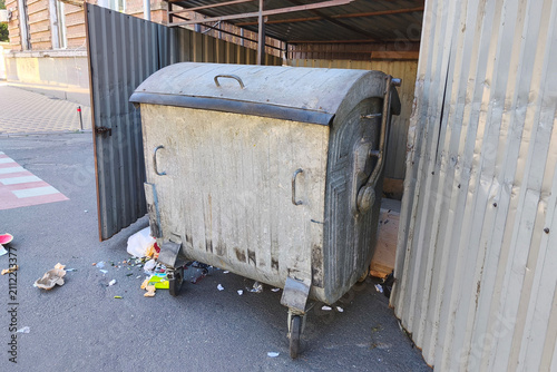 Quadro su tela Large metal garbage dumpster with scattered litter in an urban alley