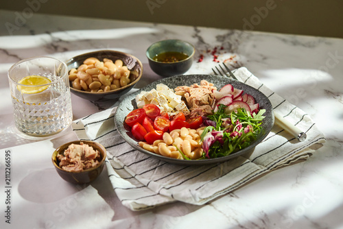 Top view of healthy tuna salad bowl with white beans, feta cheese, tomatoes, radish, arugula, and olive oil dressing on a marble surface.