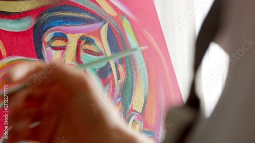 Close-up of an artist’s hand painting a colorful abstract portrait on canvas with a brush. Creative process in a bright art studio showcasing vibrant acrylic or oil painting techniques.