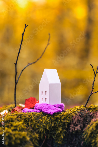 Miniature house in scarf represents passive heating concept amidst autumn setting with moss and leaves