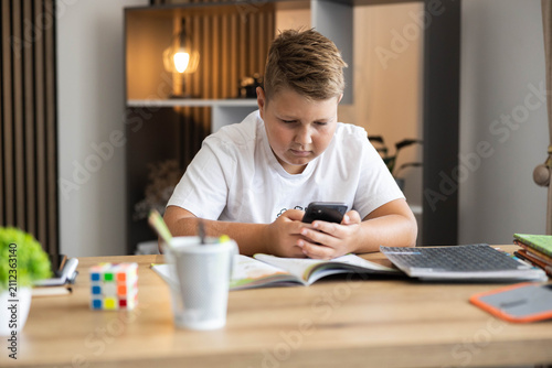 Принти на полотні Young boy absorbed in his smartphone, delaying schoolwork on a desk with an open