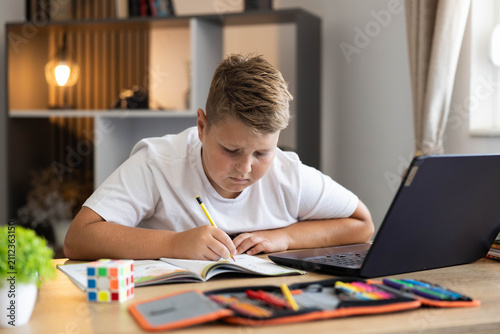 Young boy concentrating on school homework, writing in a notebook near a laptop at his desk, learning at home