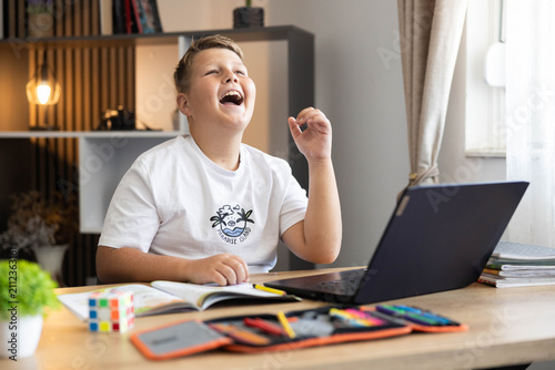 Young boy student sitting at desk, happily laughing while engaged in an online lesson or video call on a laptop