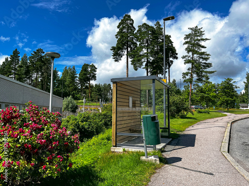 Empty Bus Stop in City Porvoo, Finland in Summer. Street, Pine Trees and Red Flowers on Lawn.