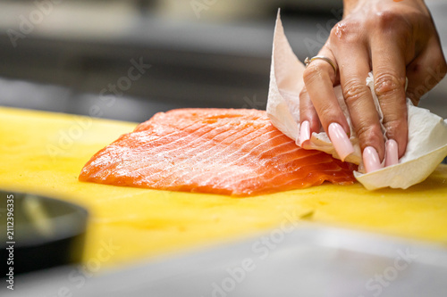Quadro su tela A chef with long pink nails pats dry a fresh raw salmon fillet on a yellow cutting board with a paper towel