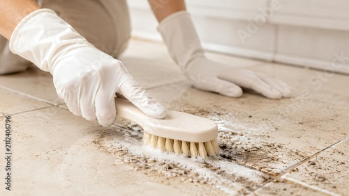 Woman scrubbing dirty tile floor with brush. Housekeeper wearing protective glove cleaning bathroom surface. Maintaining home hygiene and sanitizing living space with domestic cleaner.