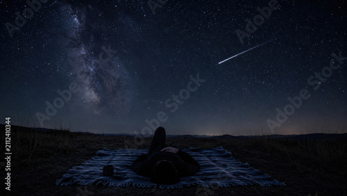 Person lies on blanket under starry sky watching bright perseid meteor shower with Milky Way shining beautifully above
