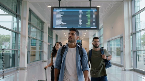 Group of friends walking through an airport terminal looking at a flight information display screen while carrying their backpacks and luggage for their vacation
