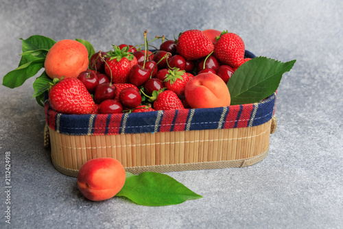 Fresh fruits peach, cherry, strawberry are arranged in a basket on a gray surface during daylight hours