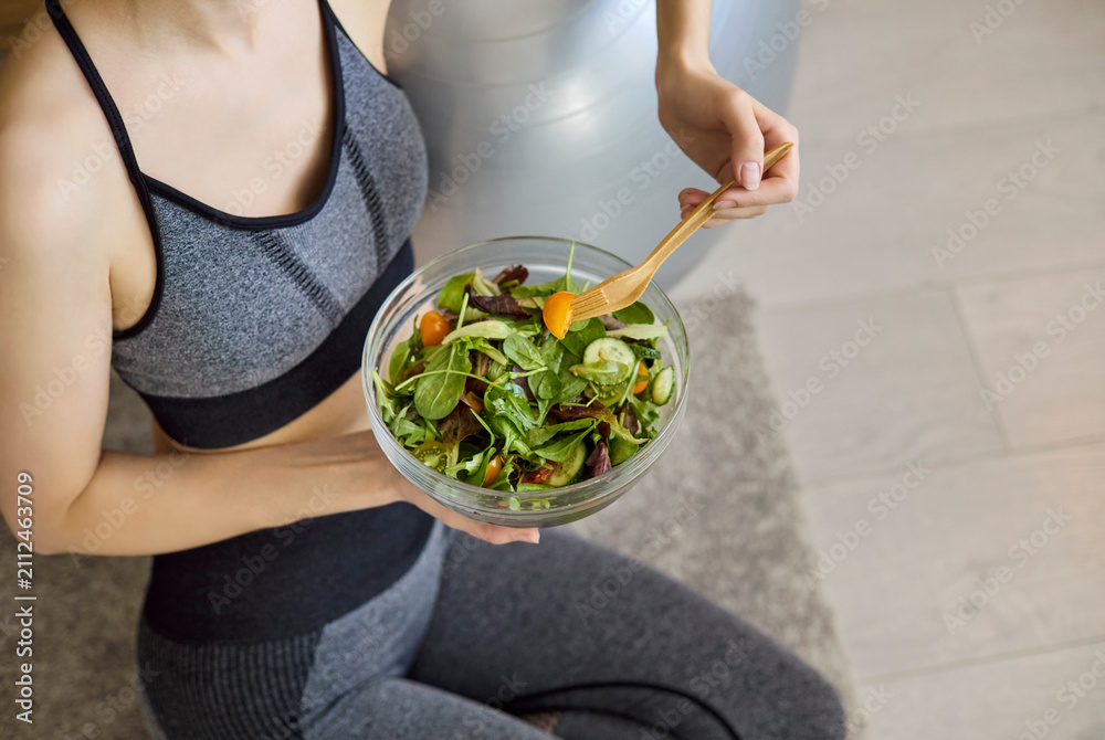 Fototapeta premium Woman eating salad after fitness at home. Athletic woman in sportswear holds a fresh salad bowl, promoting healthy diet and nutrition for wellness content. Concept of clean eating and fitness.