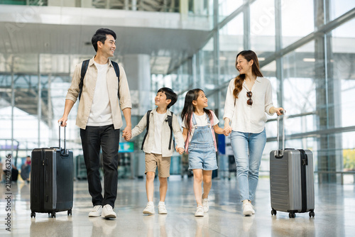 Family Vacation Awaits: A happy family of four, with parents and two children, walks hand-in-hand through a modern airport terminal, pulling their luggage.