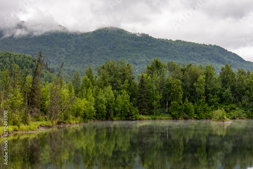 Fotografía Calm lake reflecting lush green forest and misty mountains under cloudy skies in