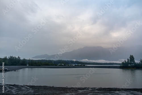 Fotografía Calm river lagoon with a gravel beach under misty mountains and a moody overcast