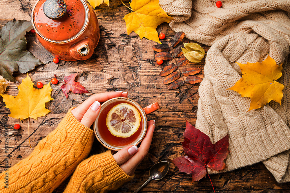 Obraz premium Cup of hot tea with lemon in woman's hands holding it over wooden autumn background with leaves and plaid Top view. Warm drink concept.
