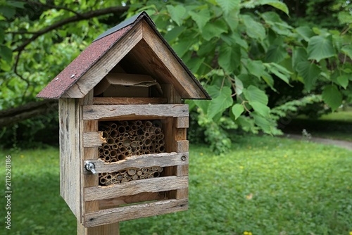Medium sized wooden insect hotel with gable type roof covered with bitumen cover, installed on wooden column in public park on cultivated lawn, summer cloudy day light. 