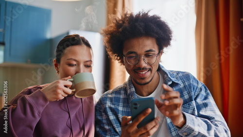 Принти на полотні Mixed race couple browsing smartphone having morning coffee in home kitchen