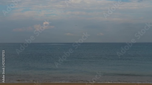 Commercial airplane descending on landing approach over calm sea and sandy beach under cloudy sky, wide static shot, daytime
