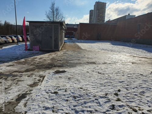 Winter urban scene featuring a small gray utility shed with closed doors positioned on a partially snow-covered paved area. A bright pink vertical flag stands near the structure. To the right, a low