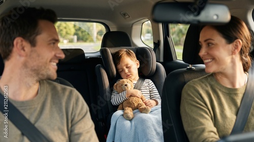 Family Road Trip Peace: A happy family enjoys a car journey, with the parents smiling at each other and their young child sleeping soundly in a car seat, clutching a teddy bear.